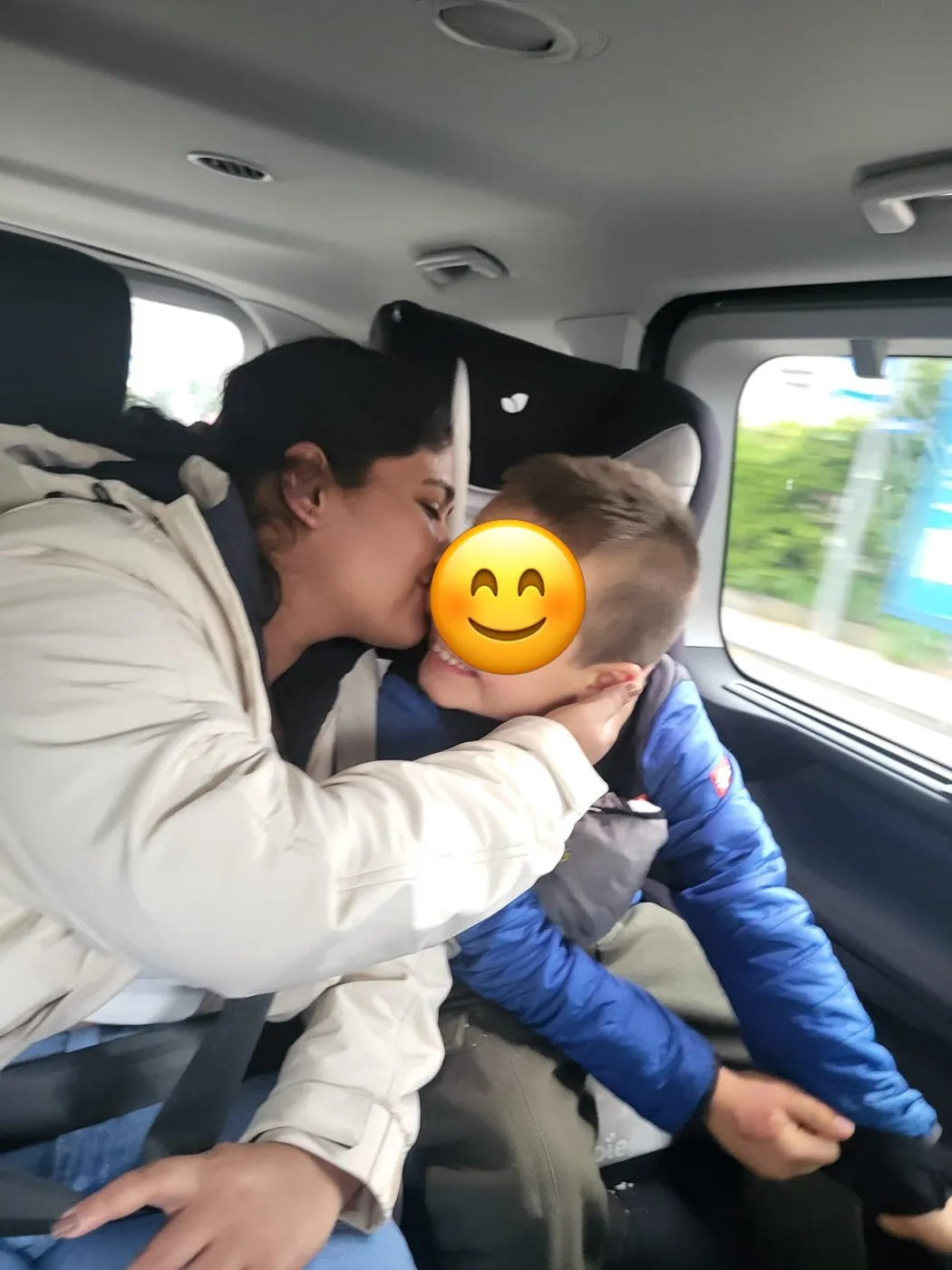 Au-pair gibt einem Kind im Auto einen Kuss auf die Wange; das Gesicht des Kindes ist mit einem Smiley abgedeckt.