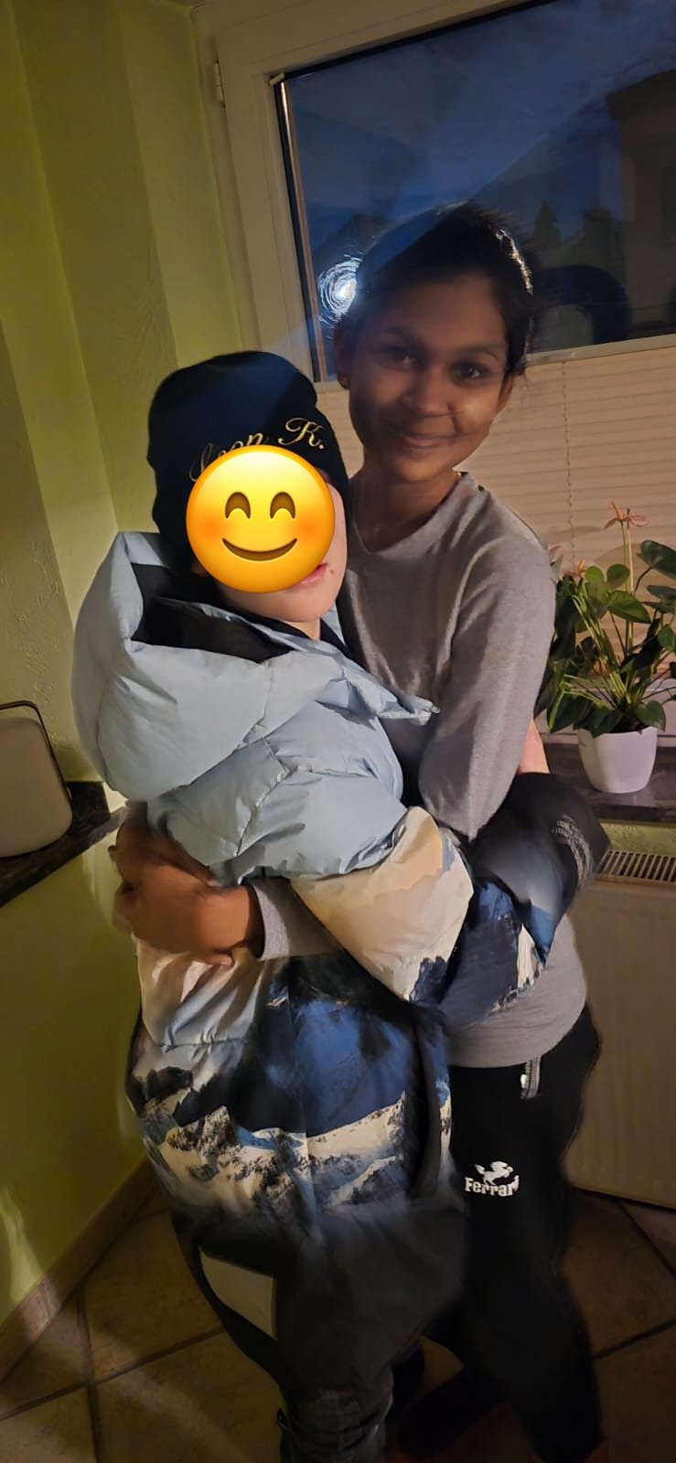 Au-pair hält ein warm angezogenes Kind auf dem Arm, beide warten auf den Schulbus; das Gesicht des Kindes ist abgedeckt.