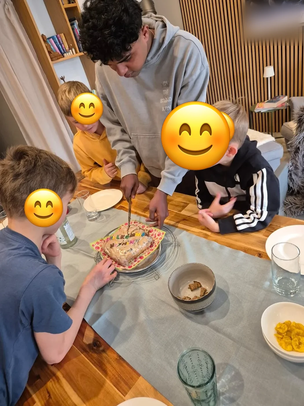 Au-pair schneidet am Esstisch einen Willkommenskuchen an, drei Kinder schauen zu; die Gesichter der Kinder sind mit Smileys abgedeckt.