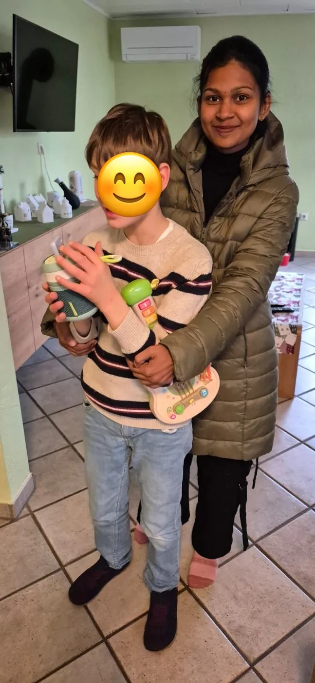 Au-pair steht hinter einem Kind und stützt es beim Spielen mit einer Spielzeuggitarre; das Gesicht des Kindes ist mit einem Smiley abgedeckt.