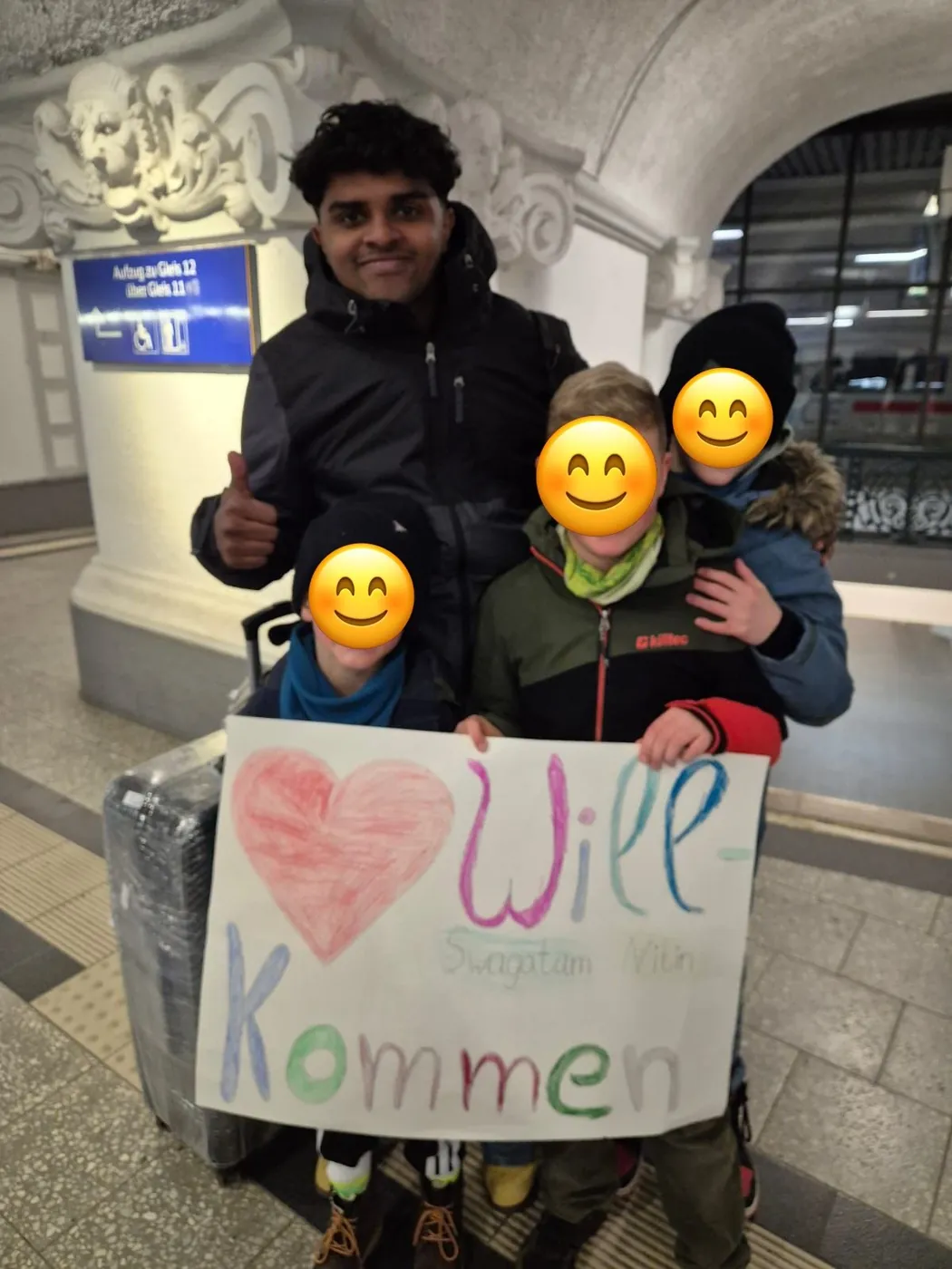 Au-pair steht mit drei Kindern am Bahnhof, die ein selbstgemaltes Willkommensschild halten; die Gesichter der Kinder sind mit Smileys abgedeckt.