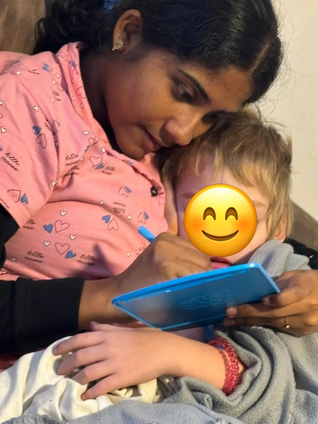 Au-pair liegt kuschelnd neben einem Kind und malt mit ihm auf einer Maltafel; das Gesicht des Kindes ist mit einem Smiley abgedeckt.