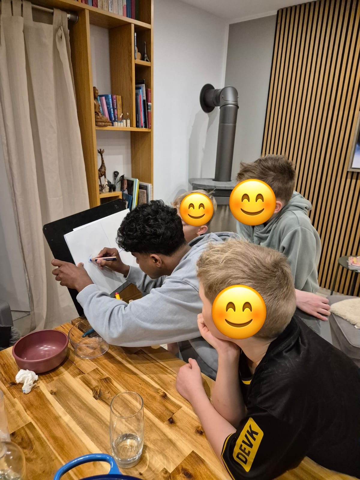 Au-pair sitzt mit drei Jungen am Esstisch und malt auf einem Whiteboard; die Gesichter der Kinder sind abgedeckt.