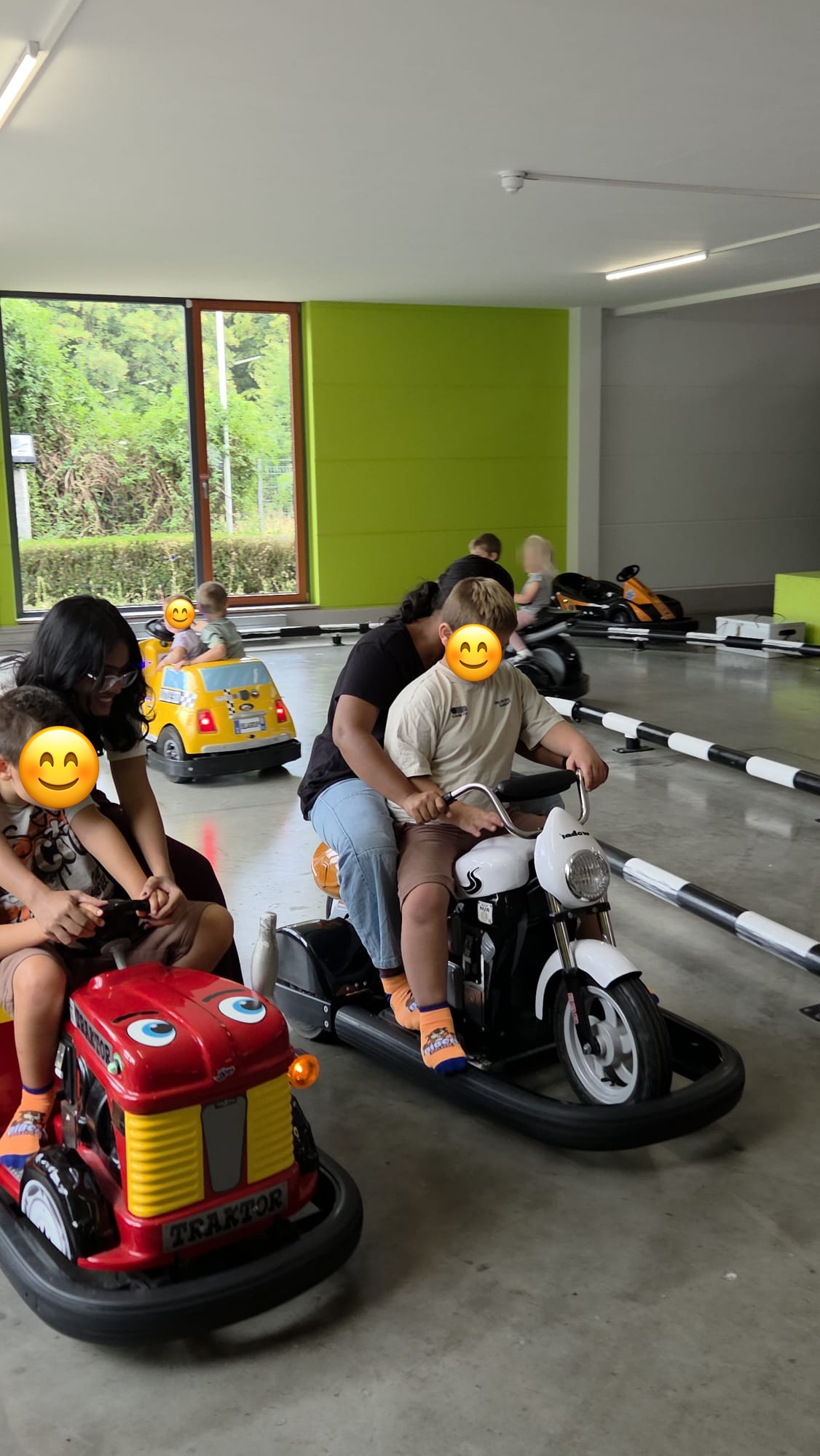 Die beiden Au-pairs mit den Kindern auf Tretfahrzeugen in der Halle; die Gesichter der Kinder sind abgedeckt.