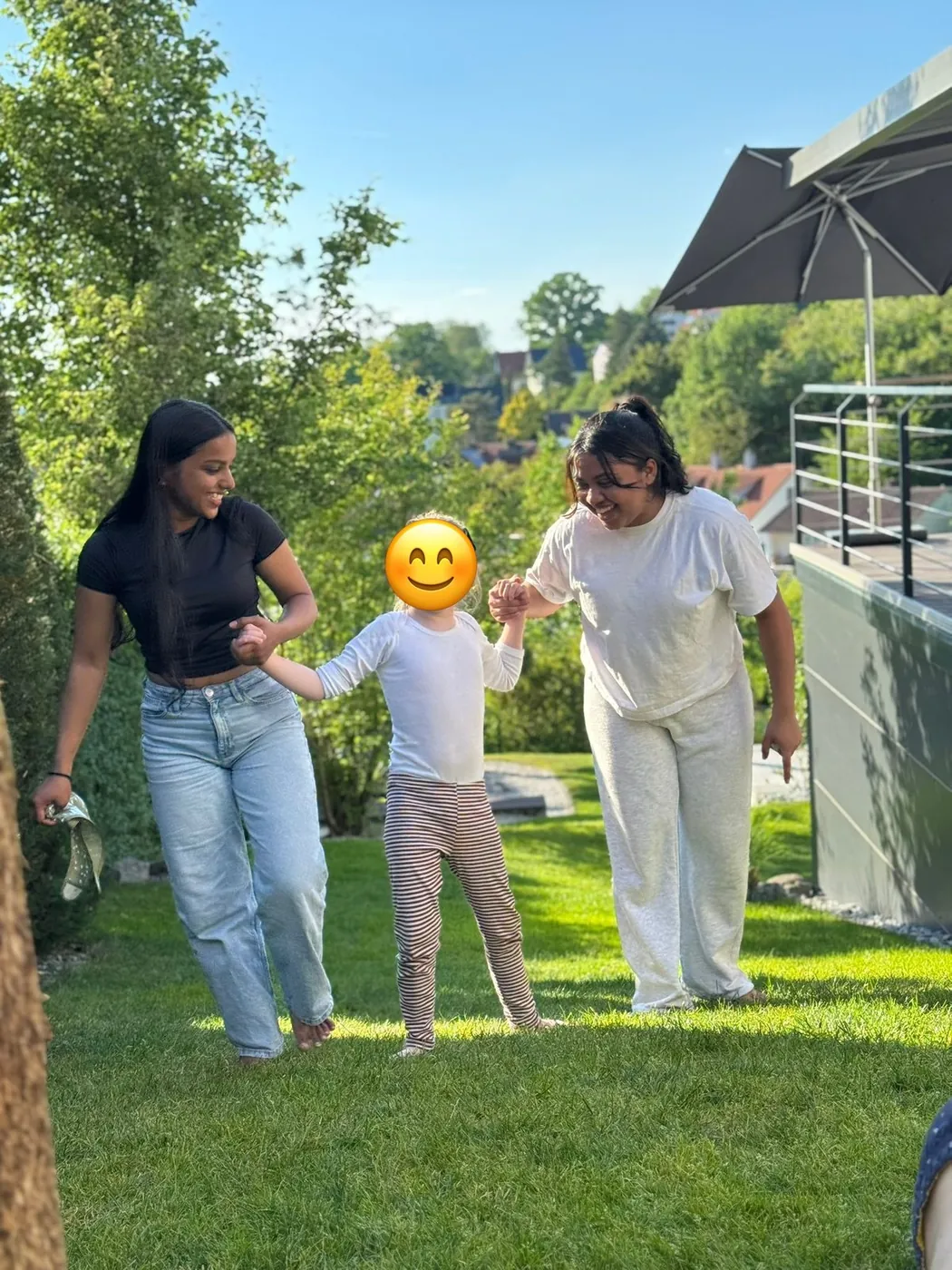 Zwei Au-pairs gehen im sonnigen Garten mit einem Kind an den Händen über die Wiese; das Gesicht des Kindes ist mit einem Smiley abgedeckt.