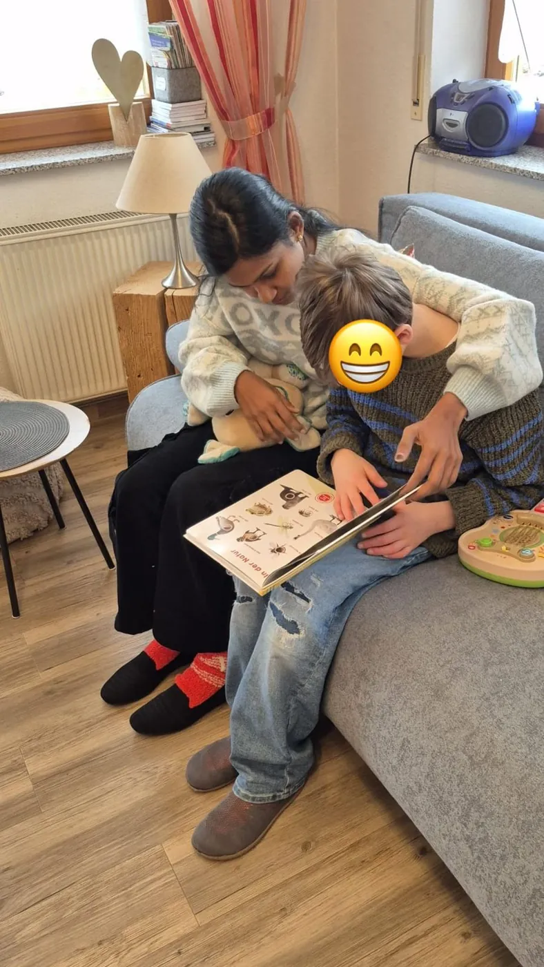 Au-pair sitzt mit einem Kind auf dem Sofa und schaut mit ihm ein Bilderbuch an; das Gesicht des Kindes ist mit einem Smiley abgedeckt.