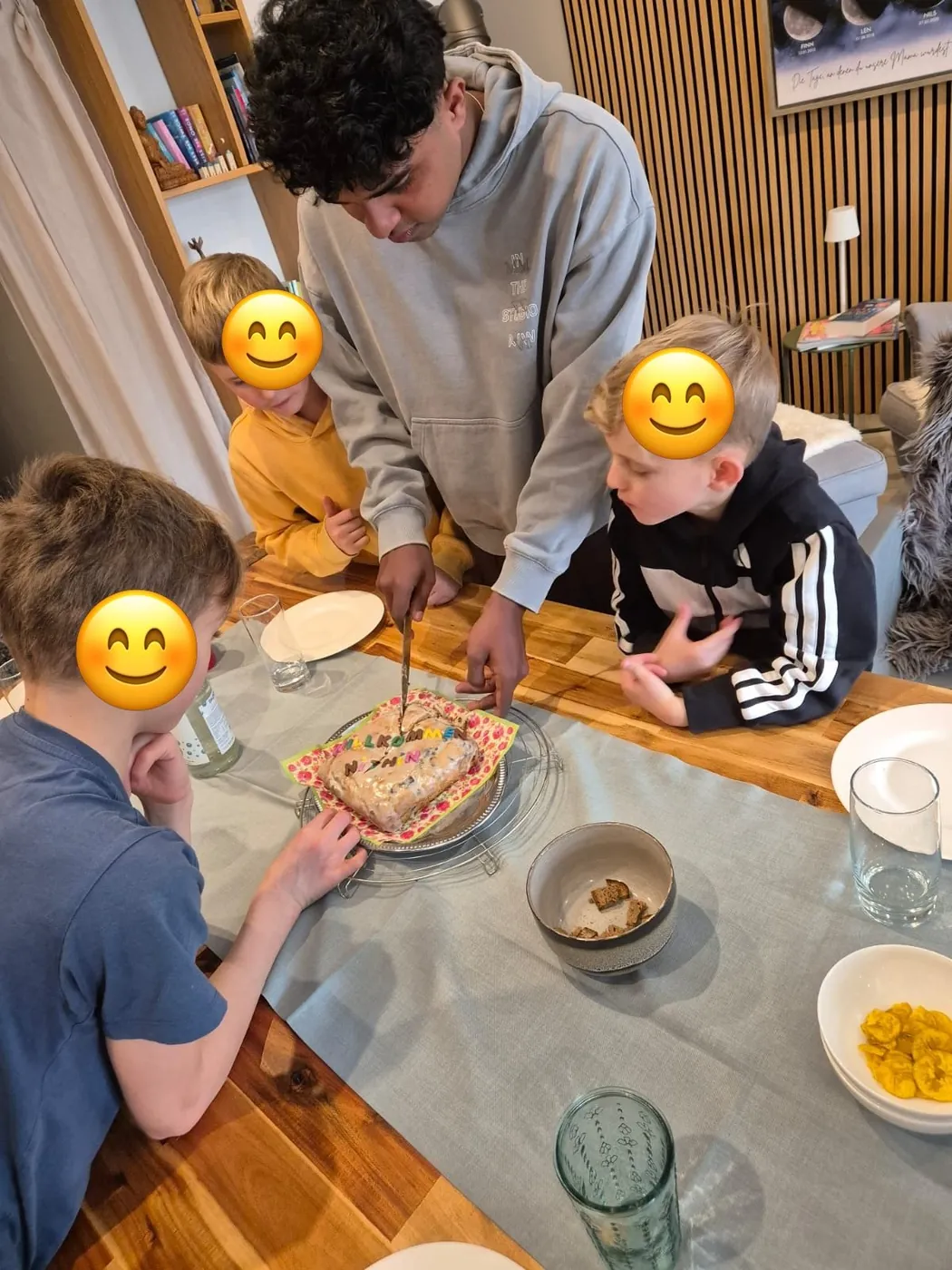 Au-pair schneidet am Esstisch einen Willkommenskuchen an, drei Kinder schauen zu; die Gesichter der Kinder sind mit Smileys abgedeckt.