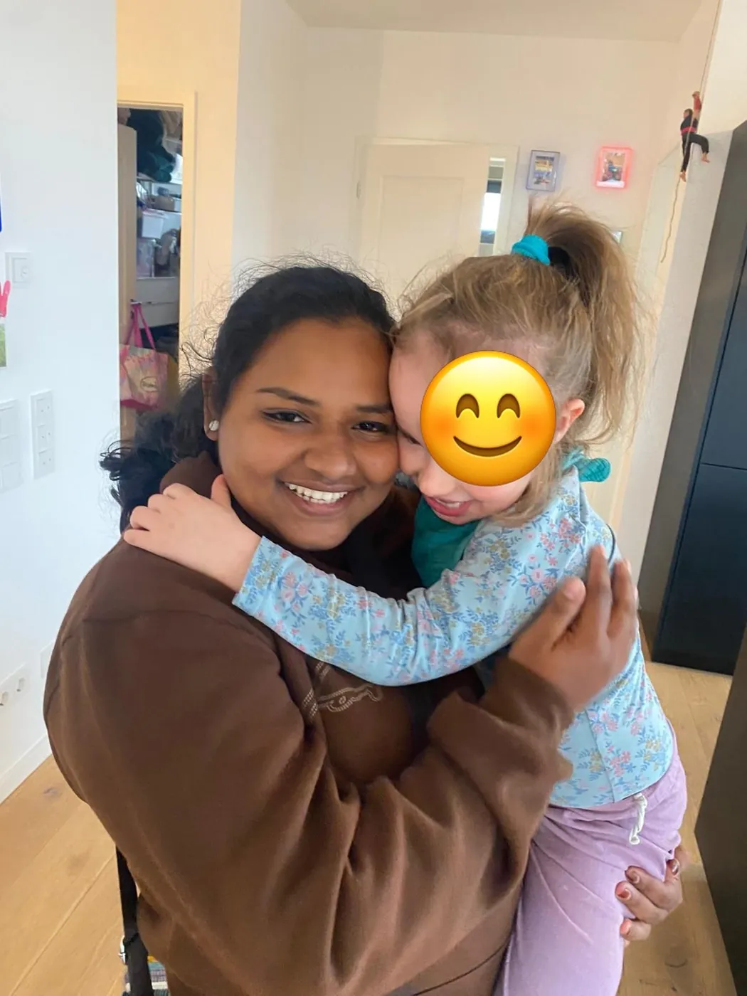 Au-pair hält lachend ein Kind auf dem Arm, das sie umarmt; das Gesicht des Kindes ist mit einem Smiley abgedeckt.