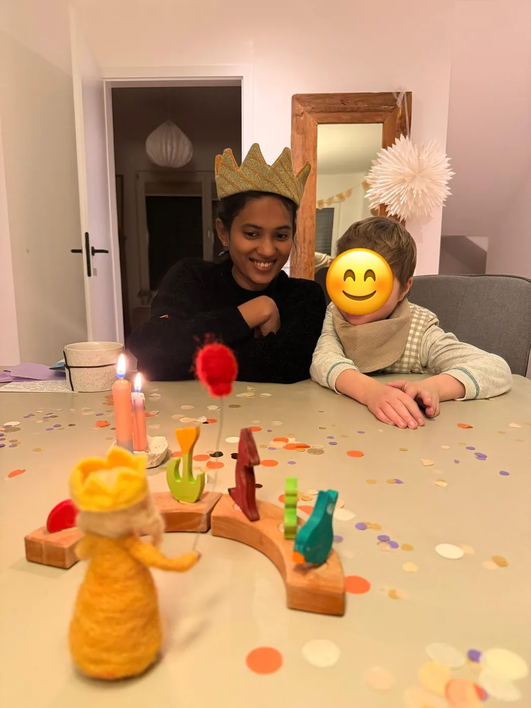 Au-pair mit Papierkrone sitzt mit einem Kind an einem festlich geschmückten Tisch mit Kerzen und Konfetti; das Gesicht des Kindes ist mit einem Smiley abgedeckt.