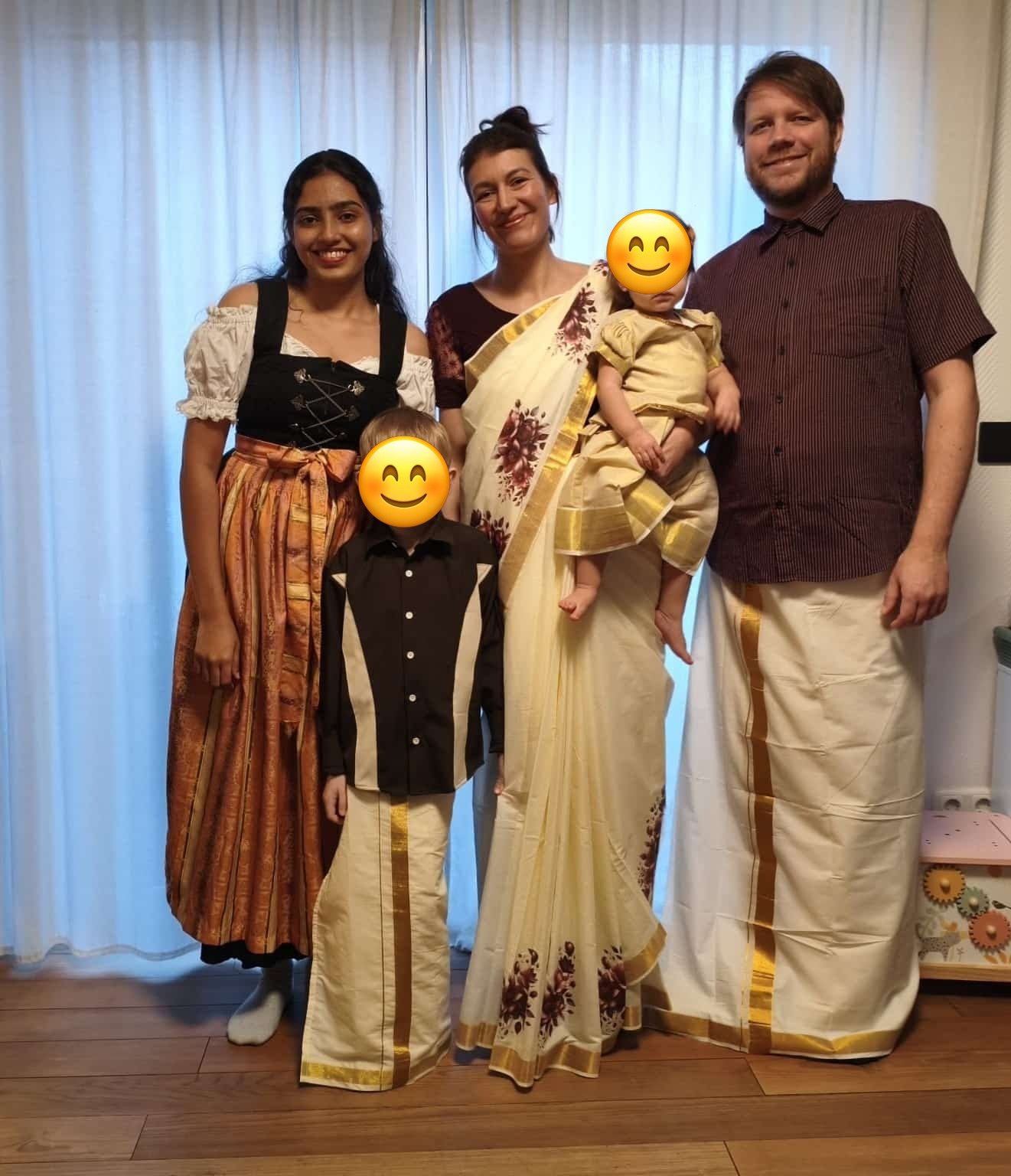 Gastfamilie und Au-pair in traditioneller Kleidung, Dirndl und Sari, nebeneinander im Wohnzimmer; die Gesichter der Kinder sind abgedeckt.