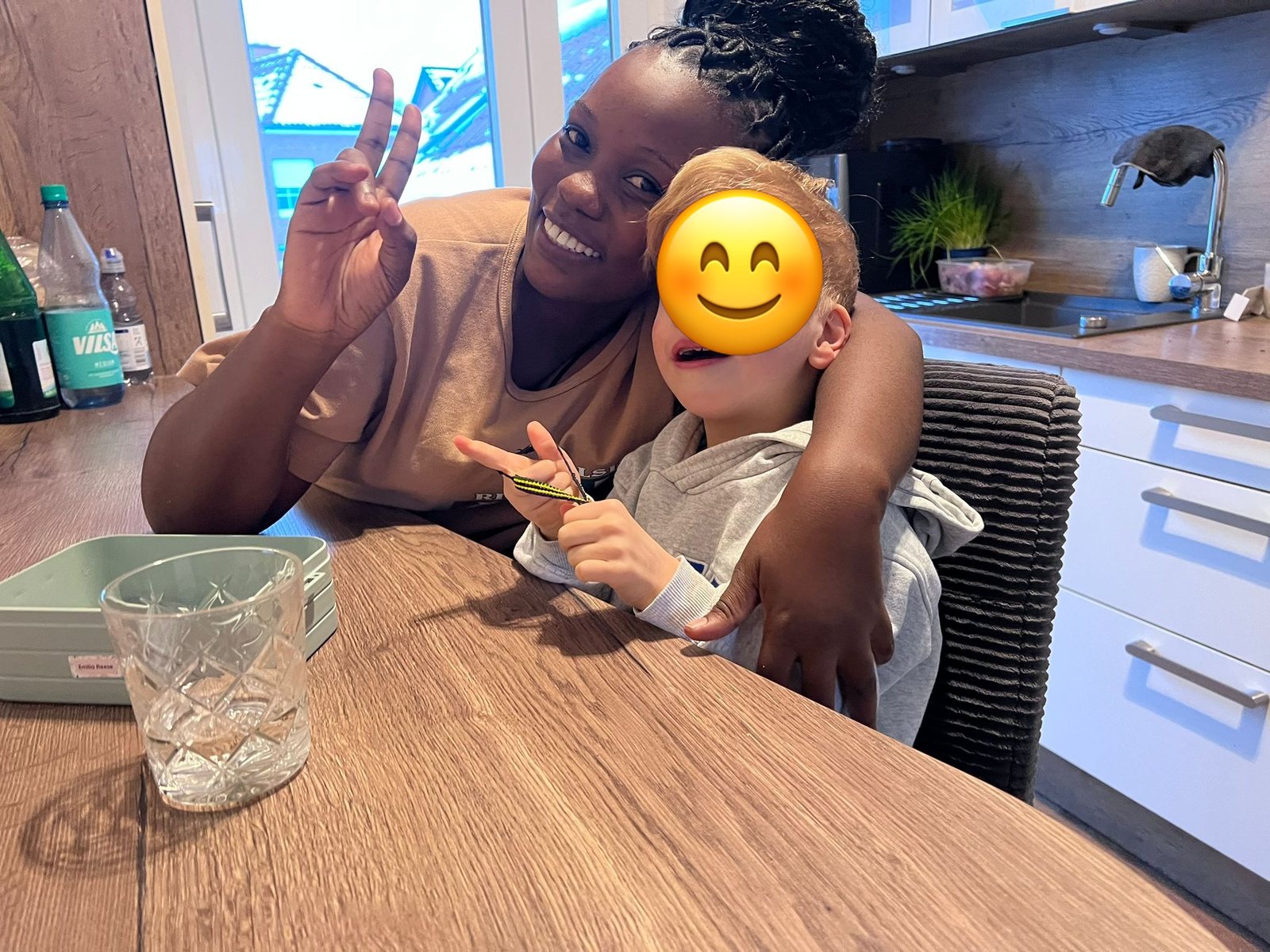 Au-pair Jailes sitzt mit einem Kind am Küchentisch, beide zeigen ein Victory-Zeichen; das Gesicht des Kindes ist abgedeckt.