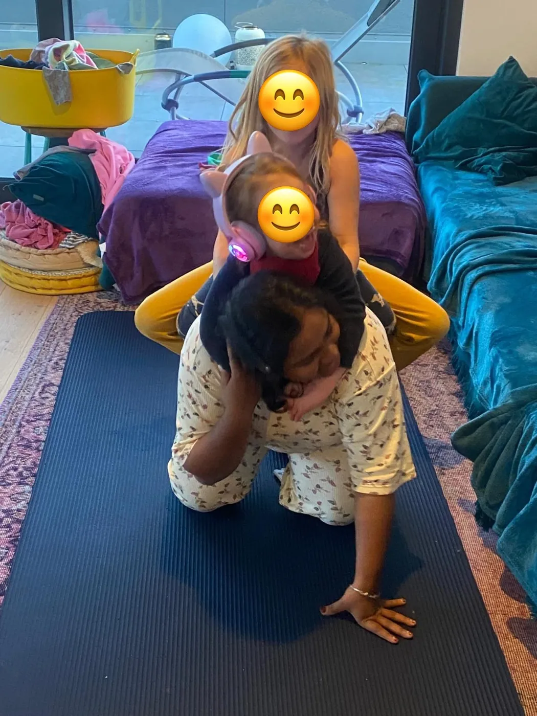 Au-pair spielt mit zwei Kindern auf einer Gymnastikmatte im Wohnzimmer; die Gesichter der Kinder sind mit Smileys abgedeckt.