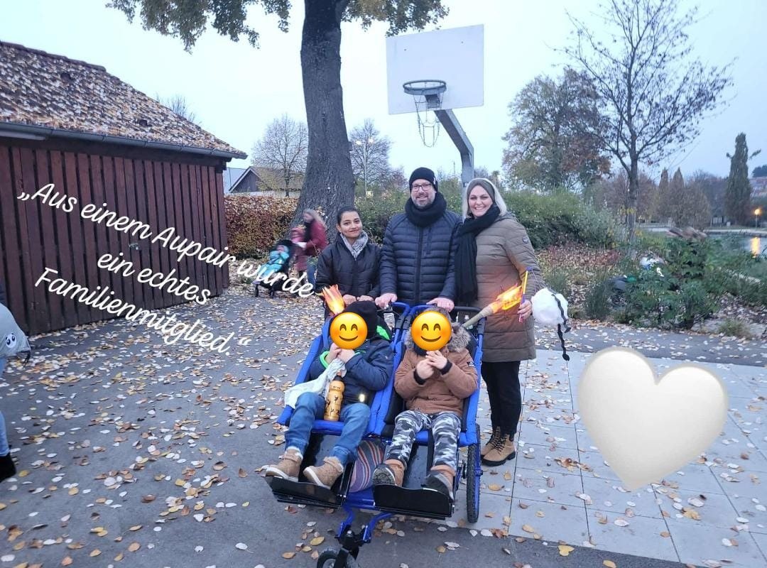Gastfamilie und Au-pair stehen im Herbst auf einem Weg hinter einem Rollstuhl-Tandem mit zwei Kindern; die Gesichter der Kinder sind abgedeckt.