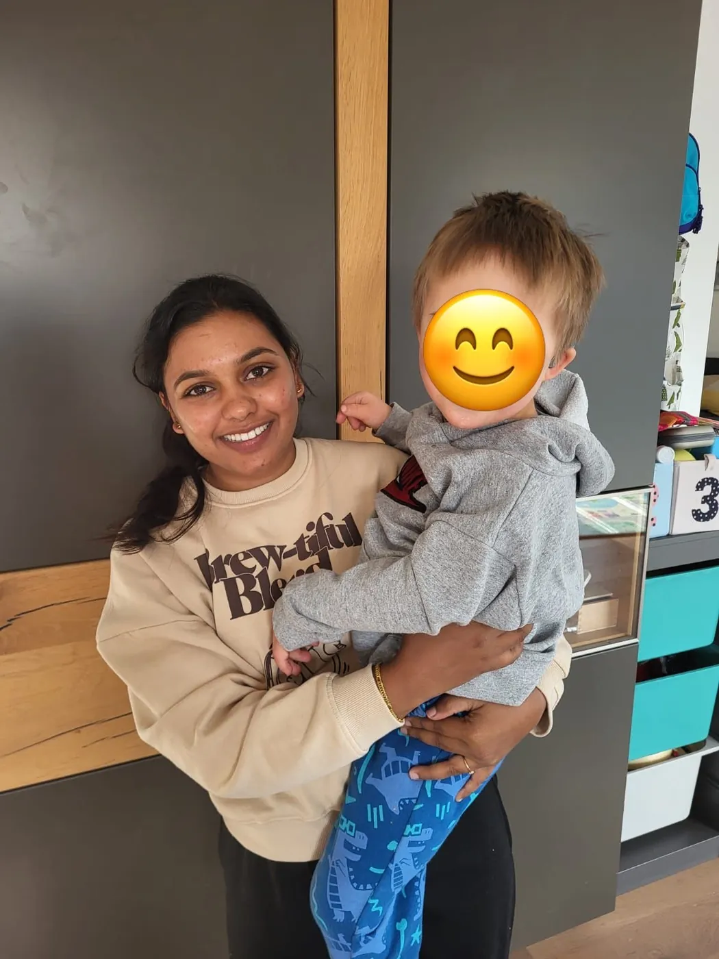 Au-pair trägt ein Kleinkind auf dem Arm in der Wohnküche; das Gesicht des Kindes ist mit einem Smiley abgedeckt.