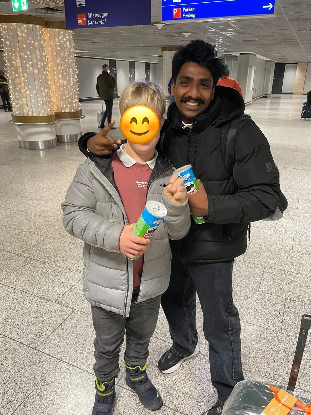 Au-pair steht mit einem Kind in einer Flughafenhalle, beide halten eine Chipsdose; das Gesicht des Kindes ist mit einem Smiley abgedeckt.