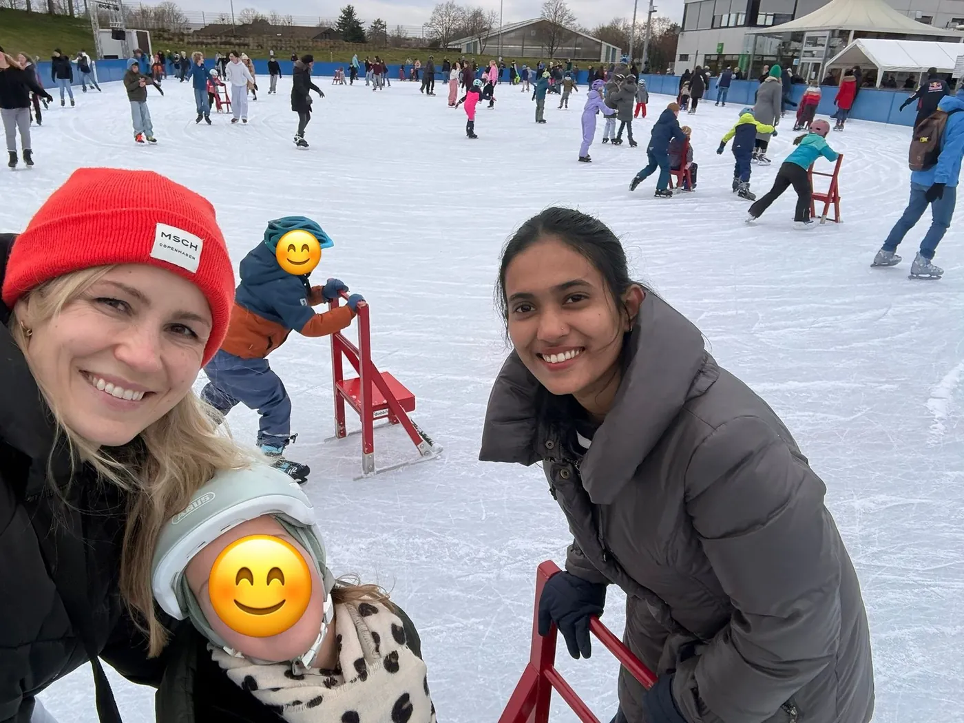 Gastmutter, Au-pair und zwei Kinder auf einer Eislaufbahn; die Gesichter der Kinder sind mit Smileys abgedeckt.