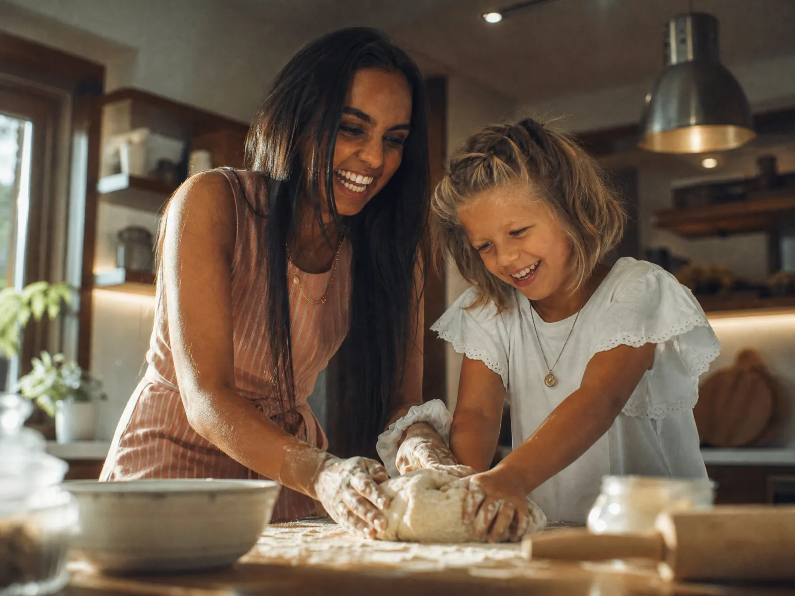 Ein Au-pair knetet gemeinsam mit einem Kind Teig auf der bemehlten Küchenarbeitsplatte, beide lachen.