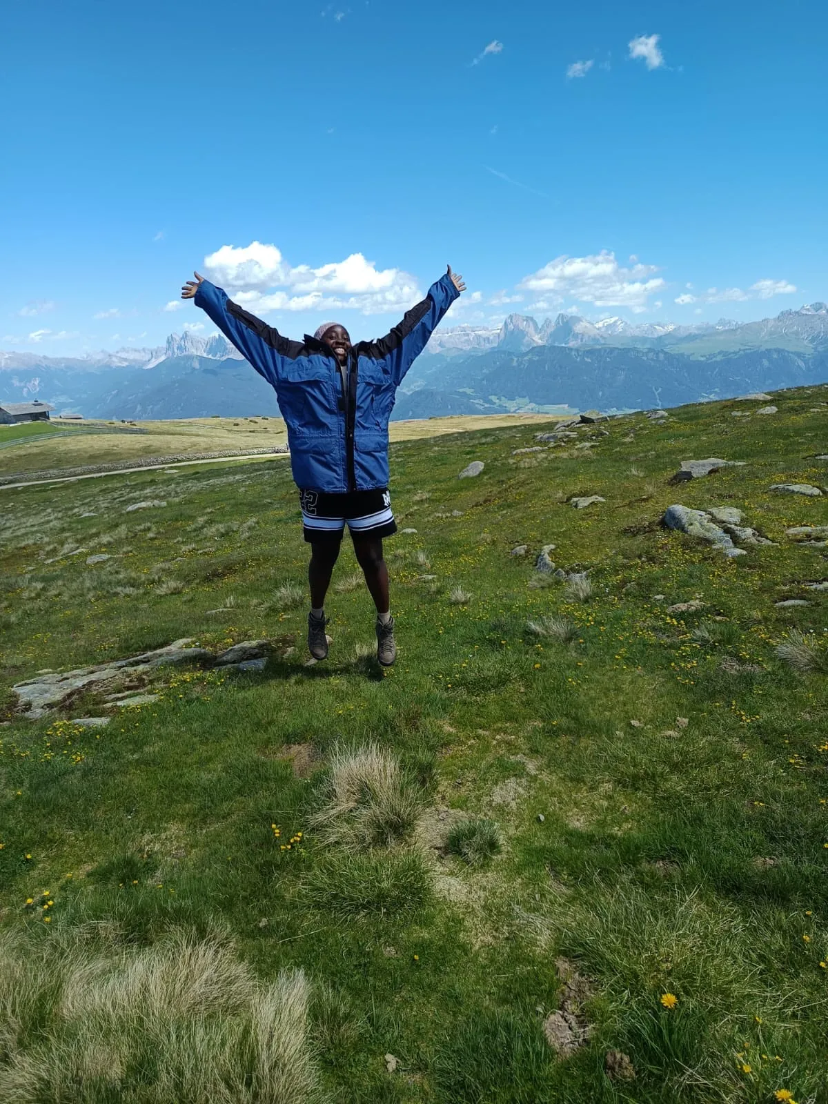 Au-pair springt mit ausgebreiteten Armen auf einer Bergwiese vor den Dolomiten in die Luft.