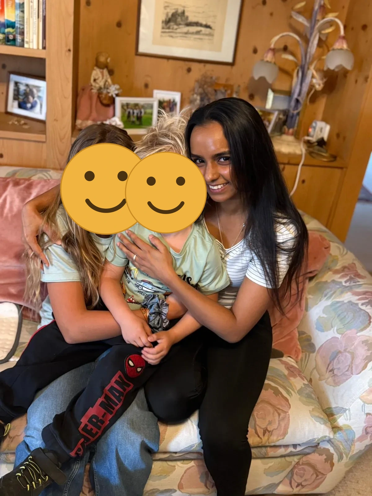 Au-pair umarmt lachend zwei blonde Kinder auf einem Sofa