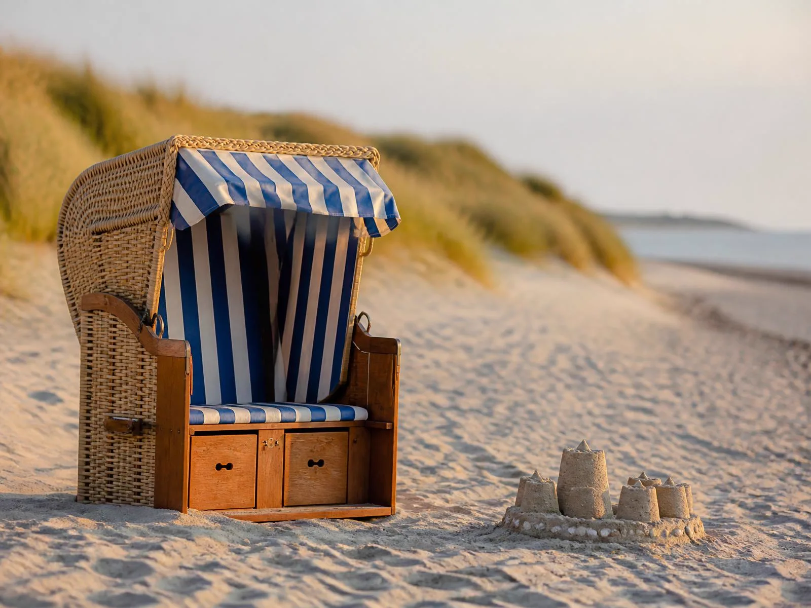 Strandkorb und eine kleine Sandburg an einem ruhigen Nordseestrand im weichen Nachmittagslicht, im Hintergrund unscharf die Dünen.