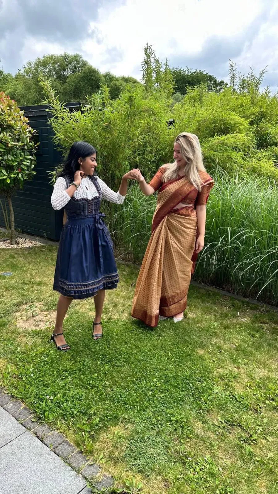 Au-pair im Dirndl und Gastmutter im Sari stehen lachend nebeneinander im Garten.