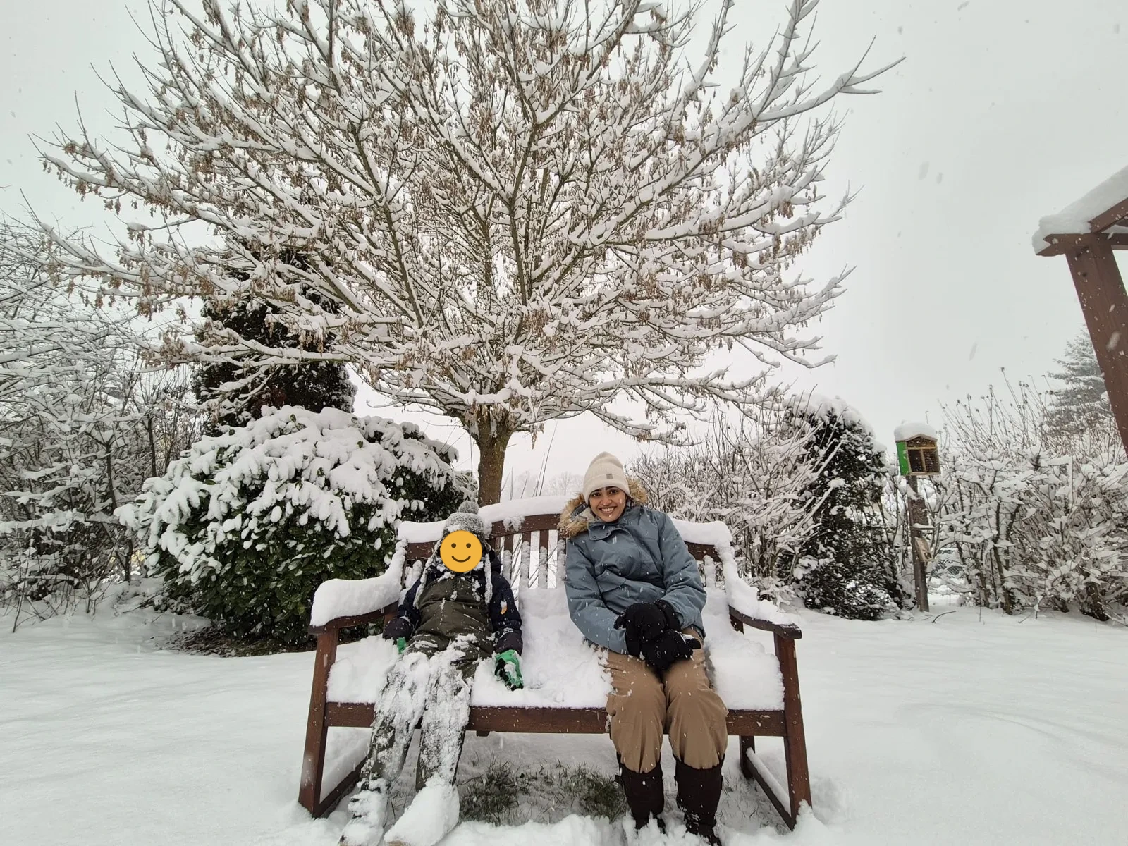 Au-pair und Kind sitzen auf einer verschneiten Gartenbank unter einem Schneebaum
