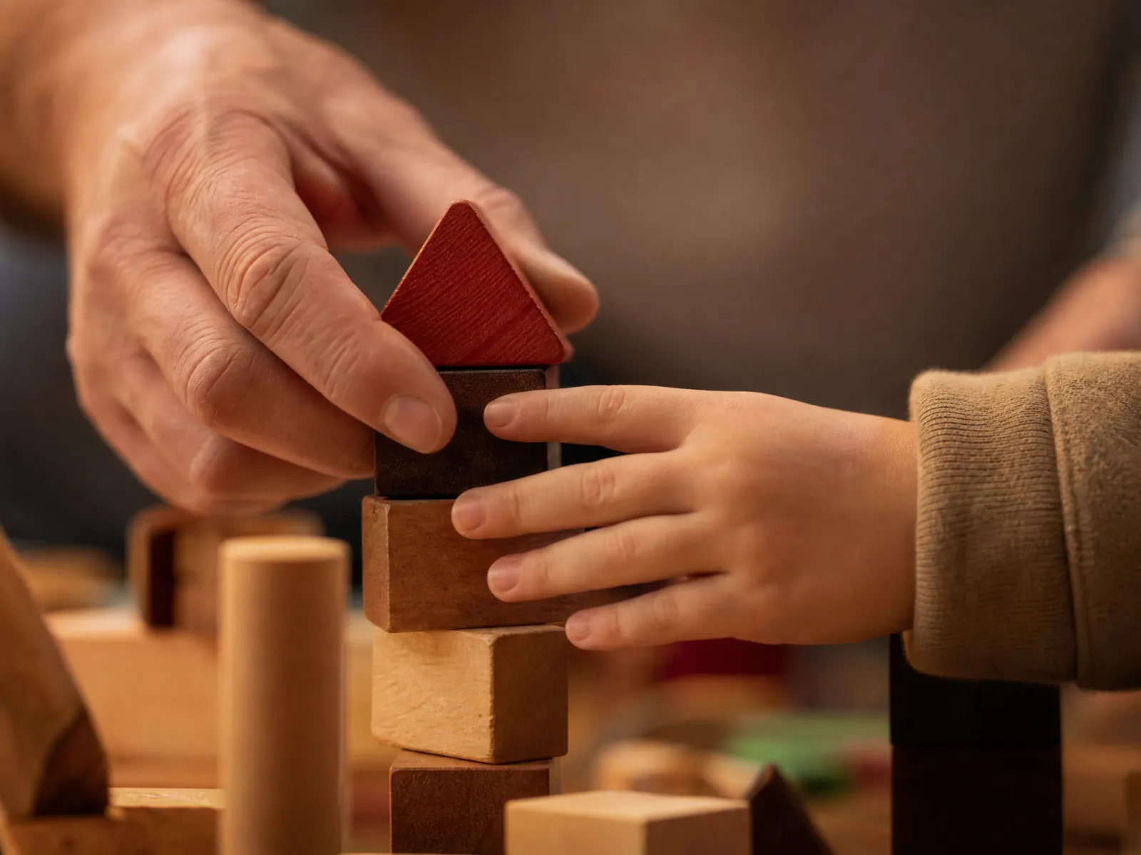 Eine Erwachsenenhand und eine Kinderhand setzen ein rotes Holzdreieck auf einen Turm aus Holzklötzen.