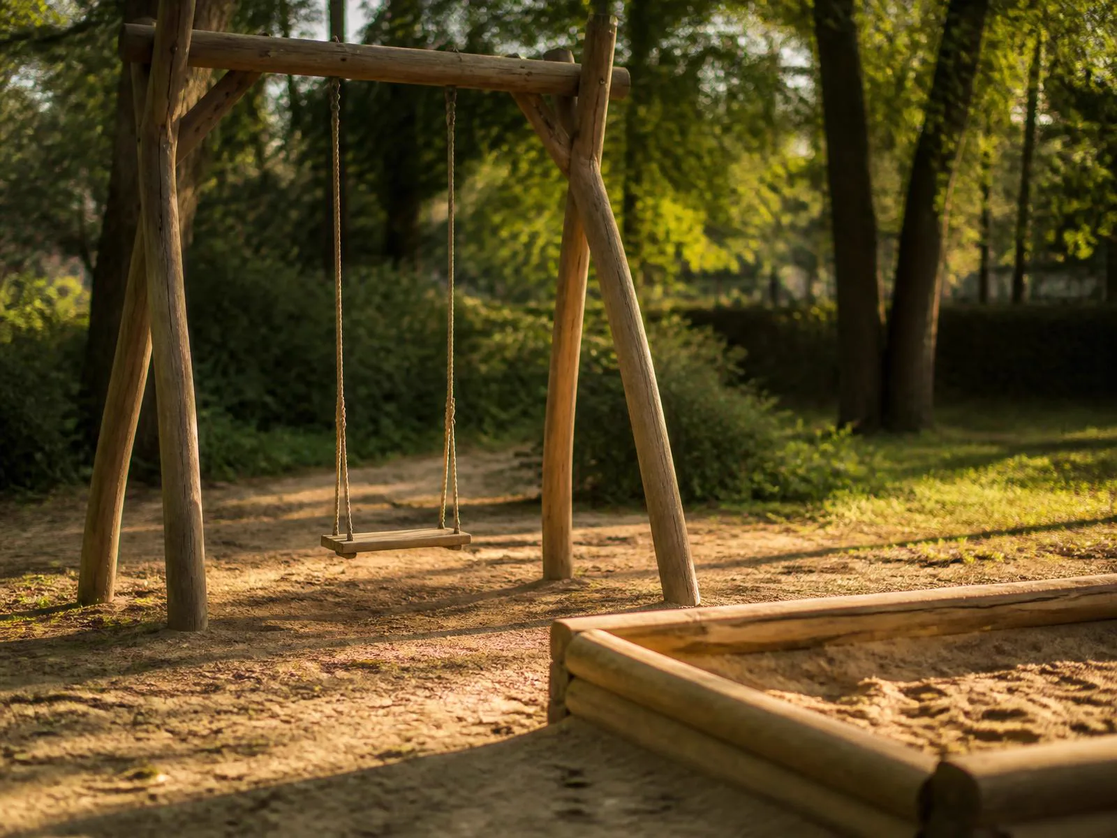 Leerer Spielplatz mit Holzschaukel und Sandkasten im warmen Nachmittagslicht, im Hintergrund Bäume.