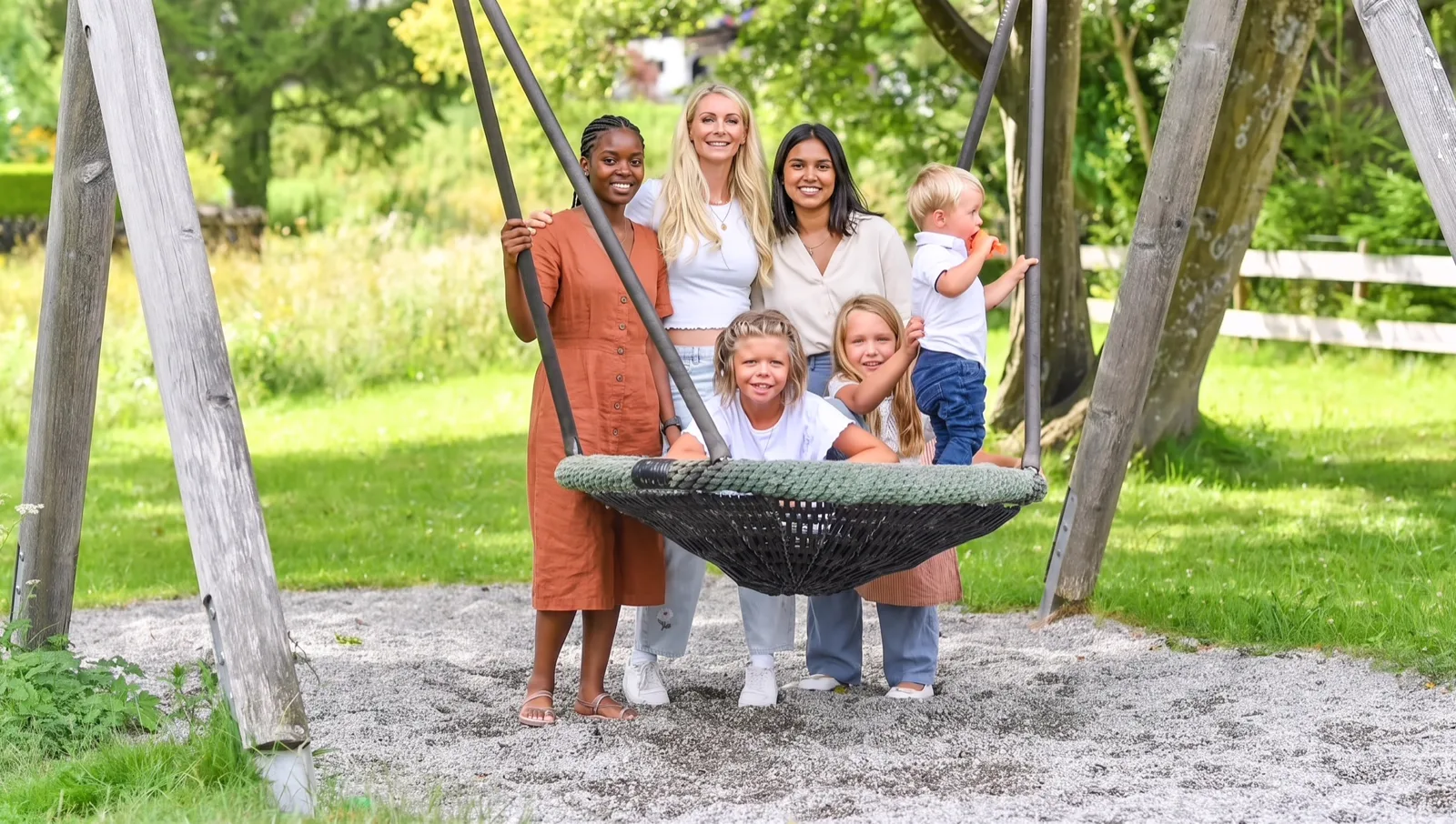 Eine Gastfamilie und drei Au-pairs stehen im Sommer lachend an einer Nestschaukel im Garten.