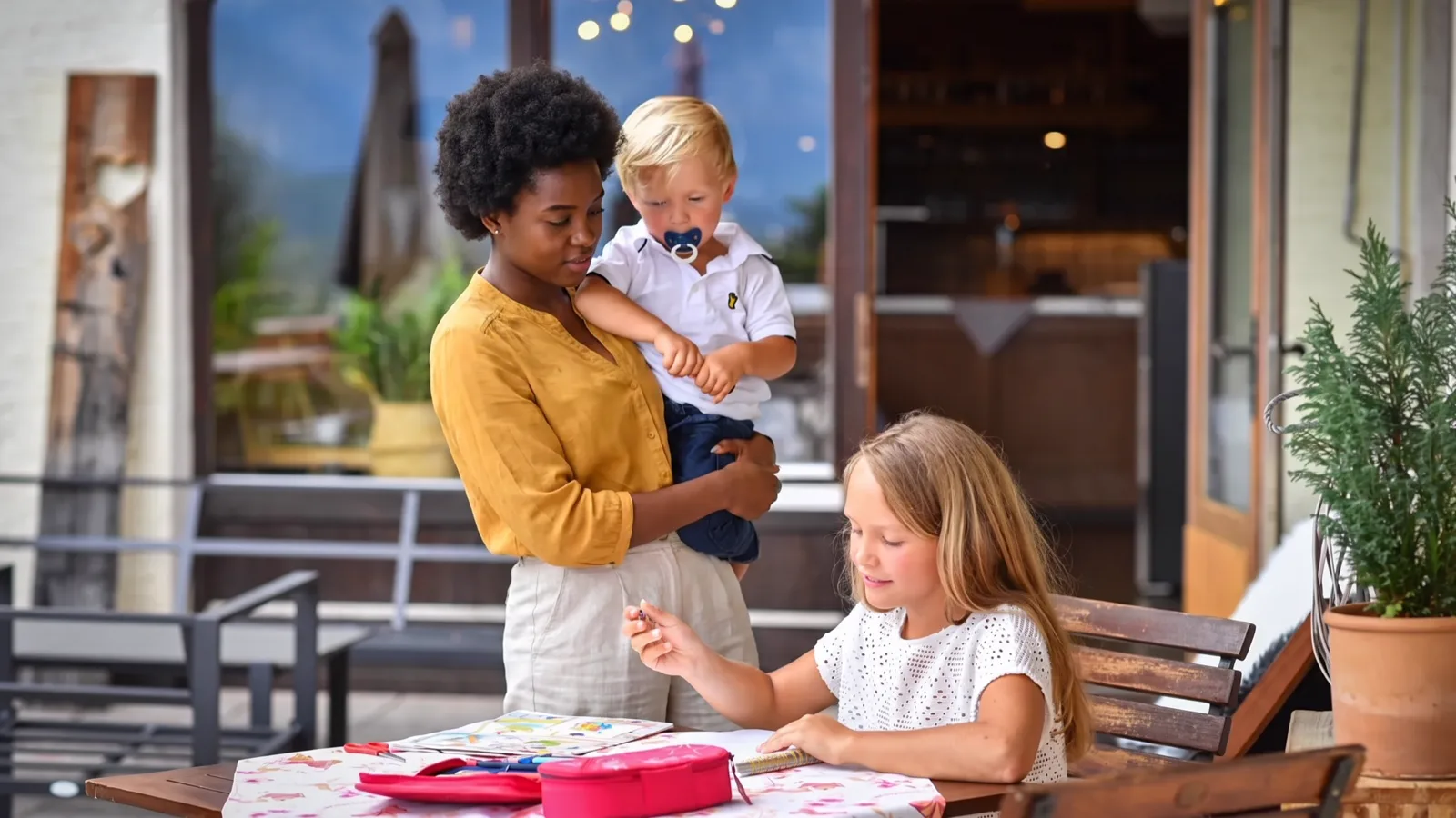 Ein Au-pair hält ein Kleinkind auf dem Arm, während ein älteres Kind am Tisch seine Aufgaben macht.