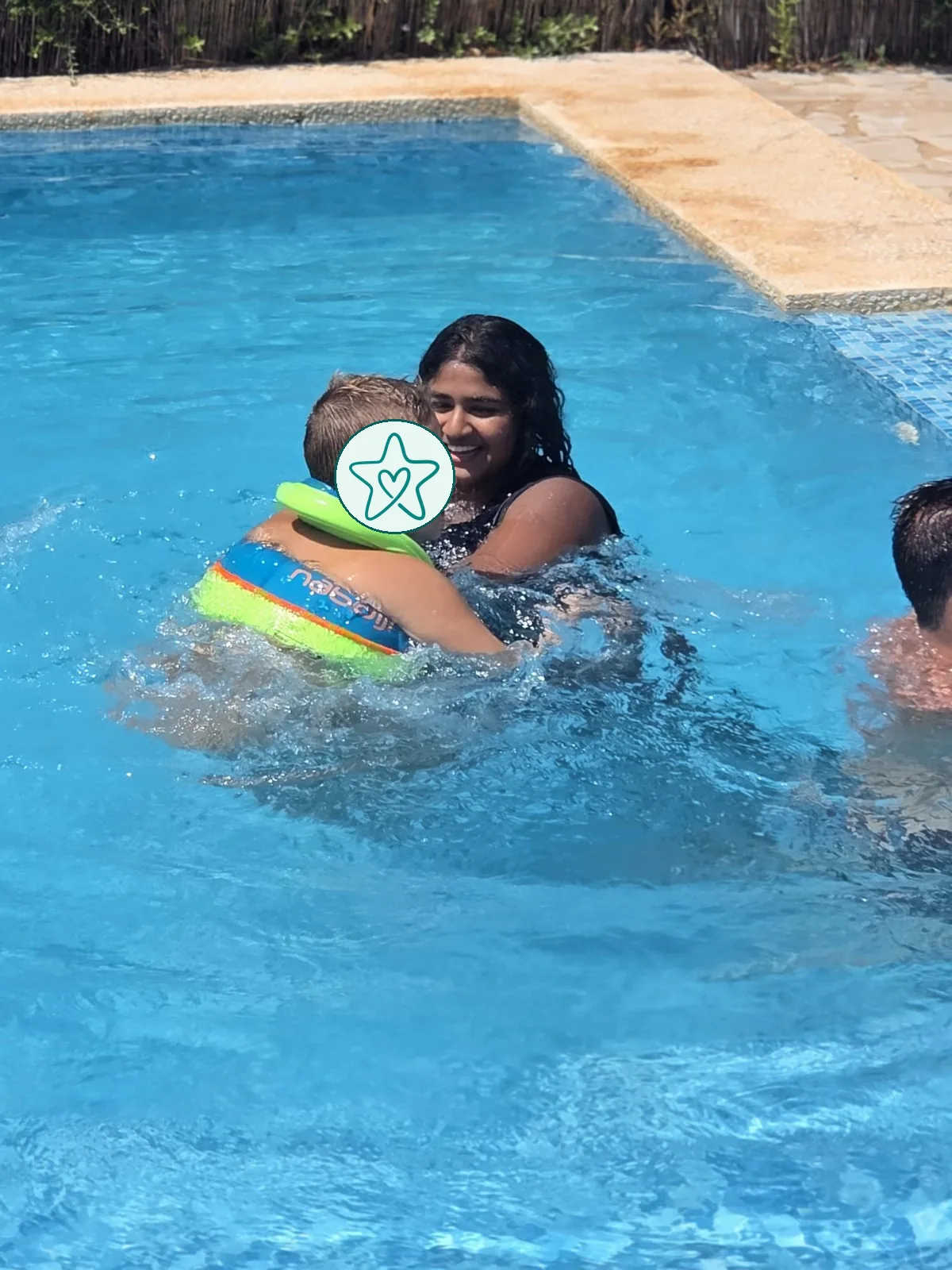 Ein Au-pair hält ein Kind mit Schwimmweste im Pool auf dem Arm, beide lachen.